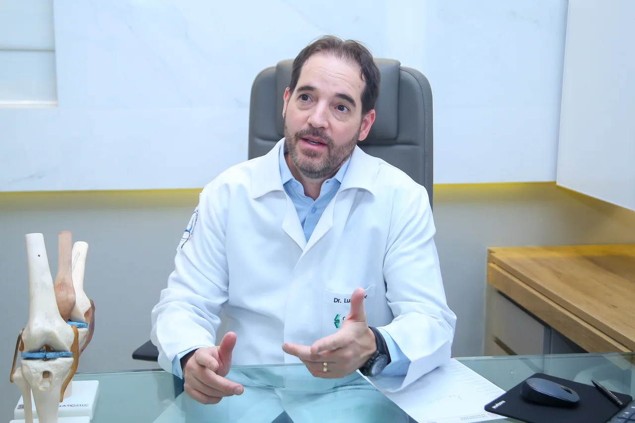 Dr. Luiz Felipe de jaleco, gesticulando ao lado de um modelo anatômico do joelho